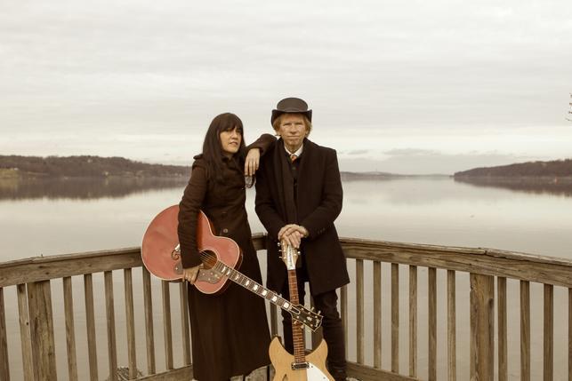 Two people holding guitars on a deck with water behind them.